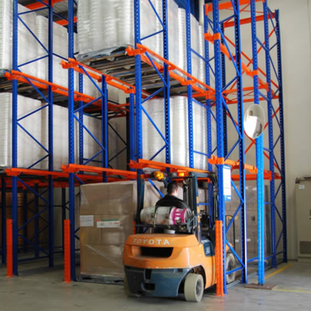 A well-lit warehouse with a drive-in racking system. Forklifts can enter the racks directly to access pallets of stacked goods.